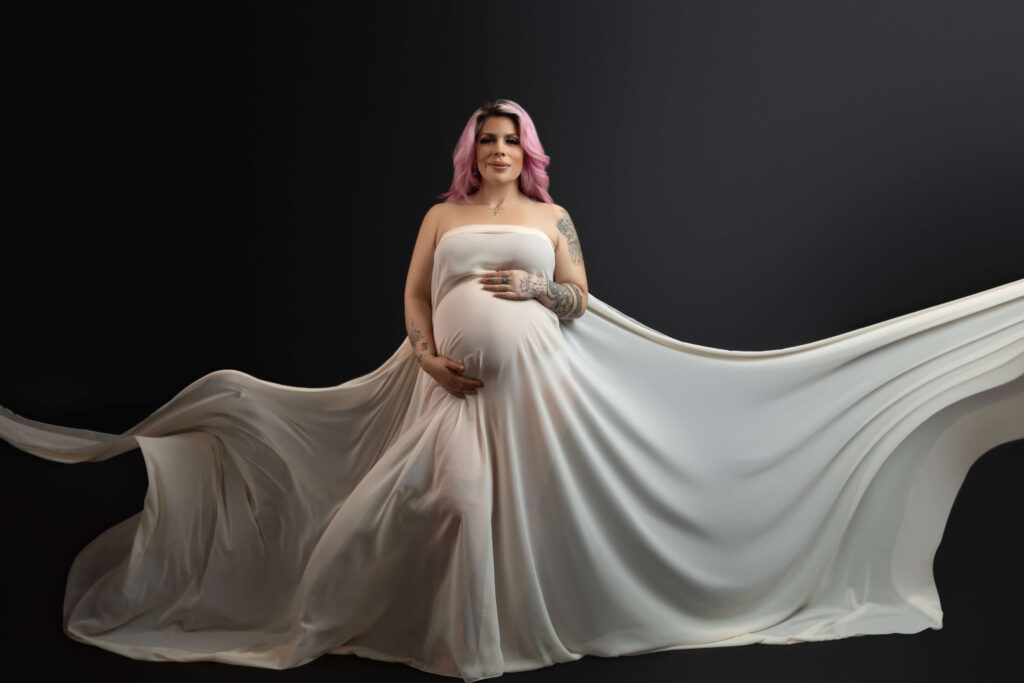 A pregnant woman with pink hair and tattoos poses in a flowing, light-colored gown against a dark background. The dress billows dramatically, creating a sense of movement and elegance.