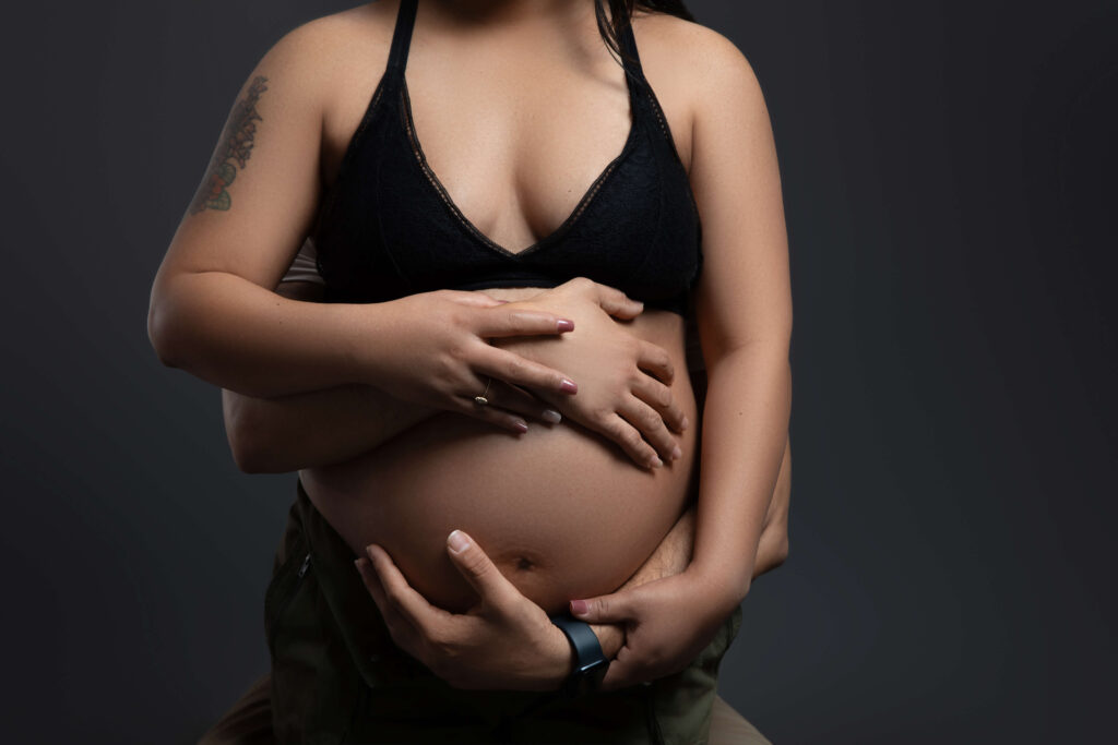 A pregnant woman in a black bra holds her bare belly, while another person gently embraces her from behind, placing their hands on her stomach. The image is set against a dark background.
