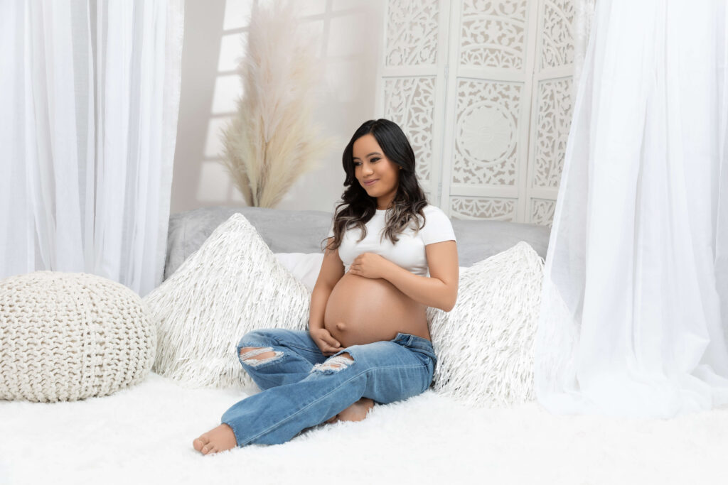 A pregnant woman sits on a bed surrounded by white pillows and drapes, wearing a white t-shirt and ripped blue jeans, smiling and holding her belly.