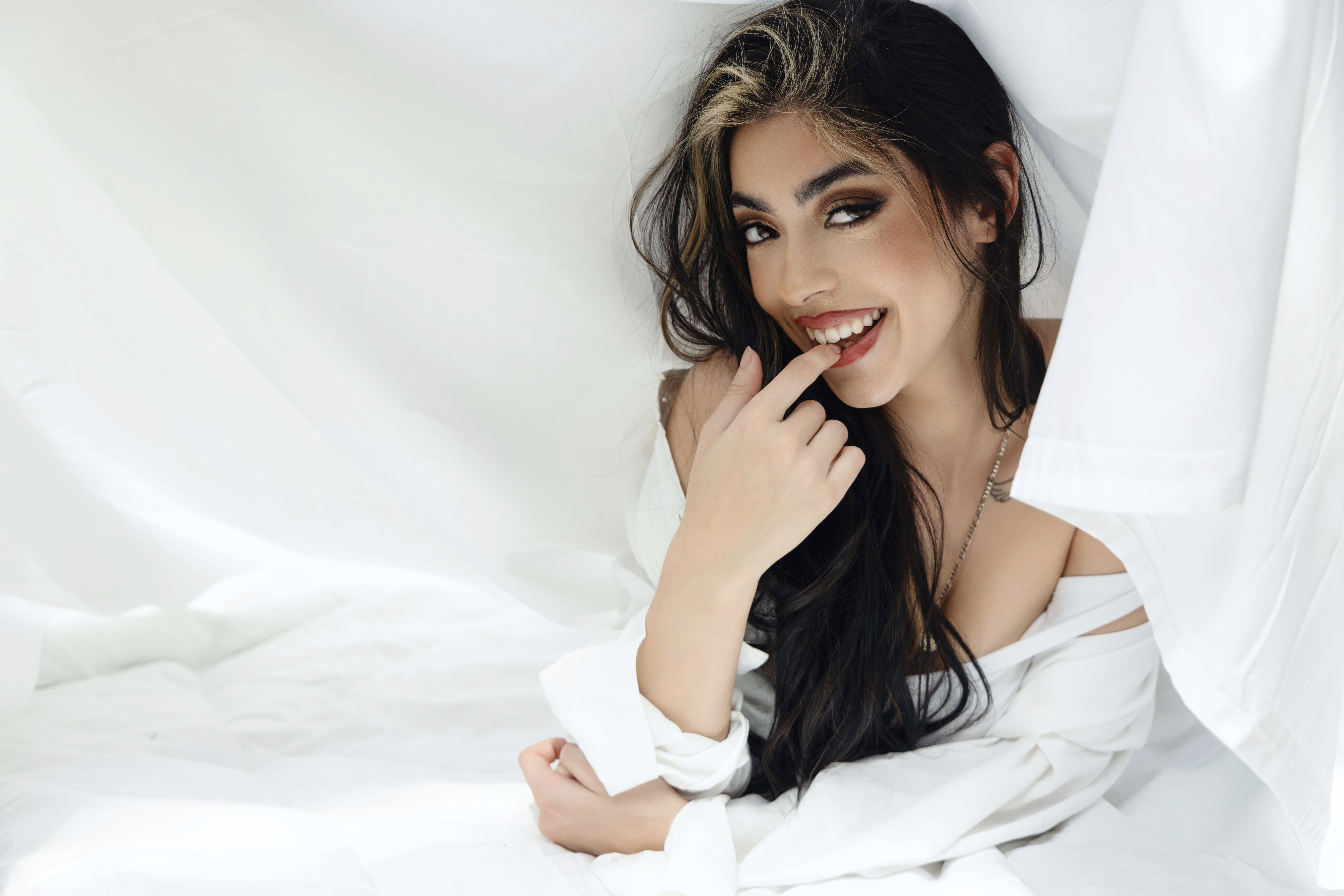 A woman with long dark hair and a streak of blonde poses playfully, touching her mouth and smiling. She wears a white shirt off her shoulders, surrounded by soft white bedding and curtains.