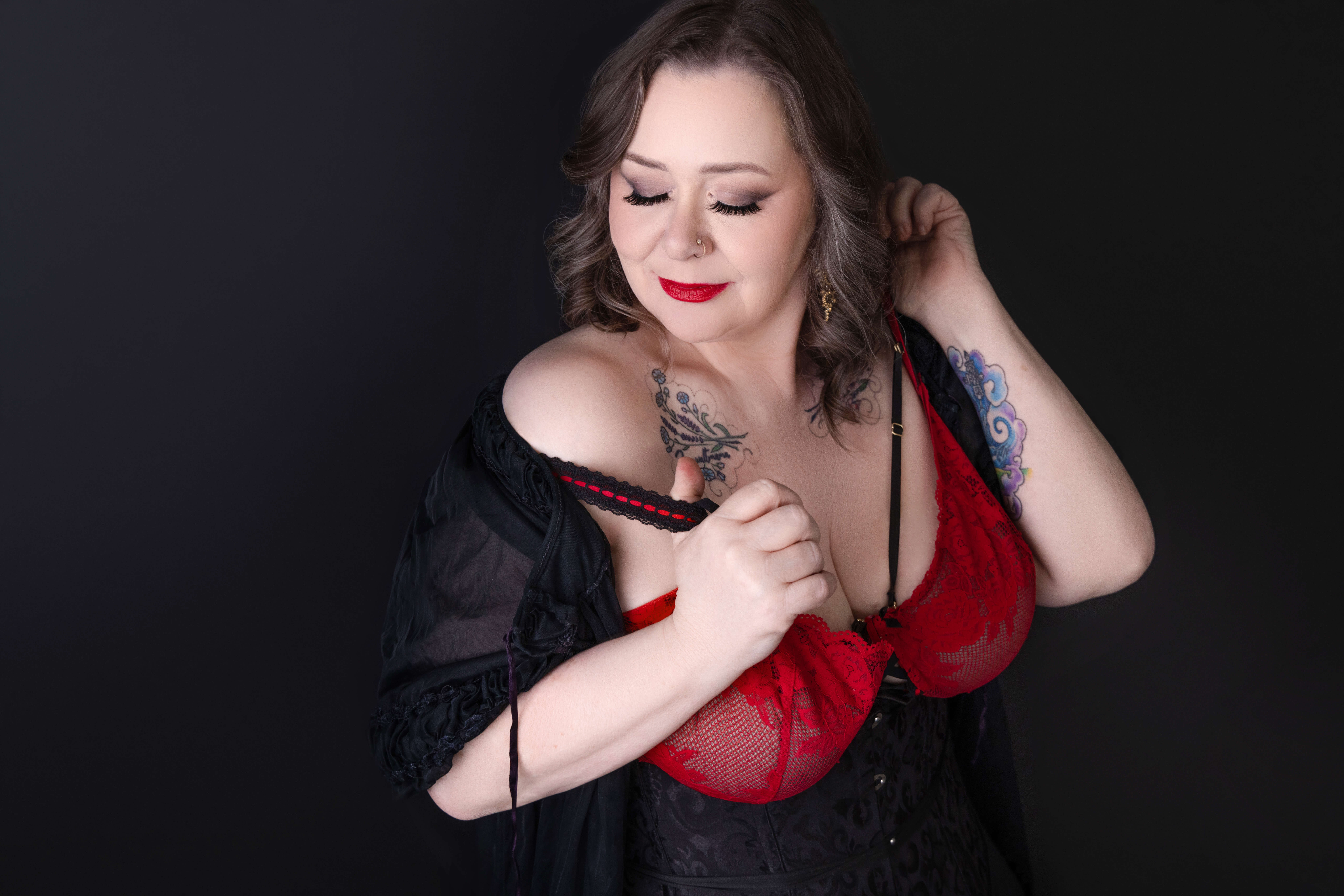 A woman with wavy brown hair wearing a red and black lace outfit looks downward, gently pulling off one strap and touching her hair. She has tattoos on her shoulder and arm, with a soft, dark background.