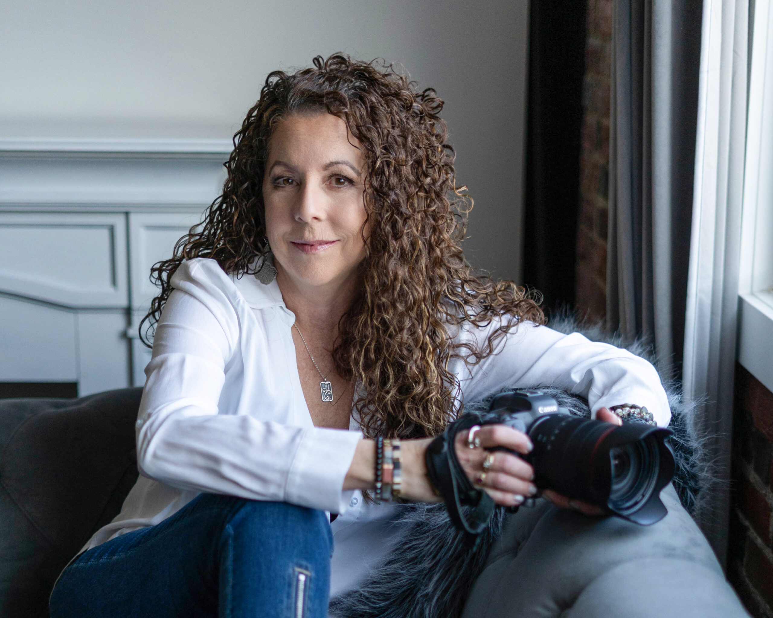 A woman with curly brown hair sits on a gray couch by a window, wearing a white blouse and jeans, holding a camera and looking at the camera with a slight smile.