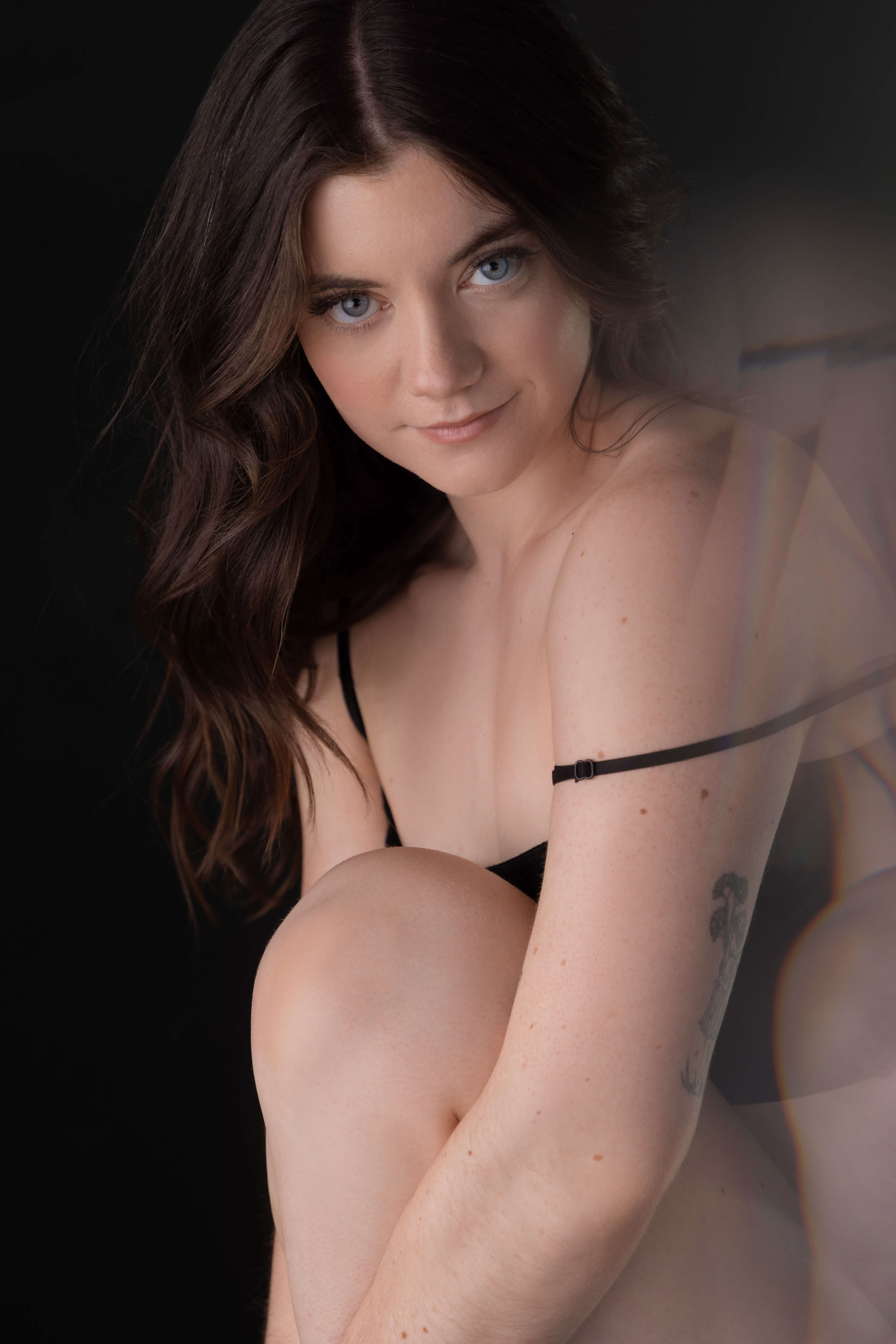 Woman seated in black lingerie on foot stool with arms wrapped around leg looking at camera during a private boudoir session in Wenatchee.